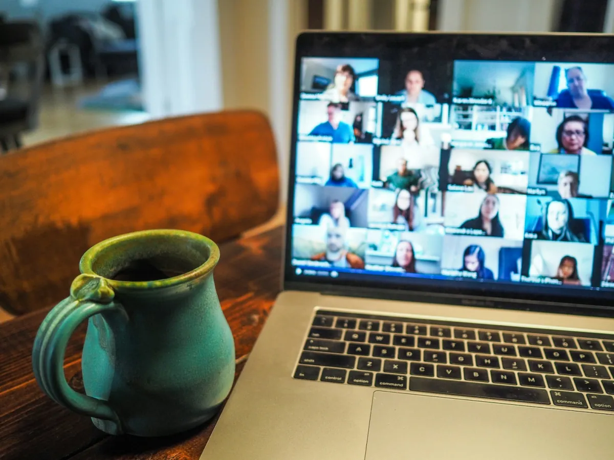 A laptop on a wooden table showing a video call with a grid of colleagues, beside a green ceramic mug of coffee